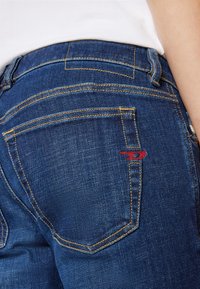 Vaqueros de denim azul con costuras amarillas, que presentan un pequeño logo rojo en el bolsillo trasero, diseño clásico de cinco bolsillos y una cintura de altura media.