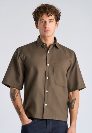 Młody mężczyzna z kręconymi włosami, ubrany w brązową koszulę na krótki rękaw zapinaną na guziki oraz niebieskie dżinsy, pokazujący tatuaże na obu przedramionach.