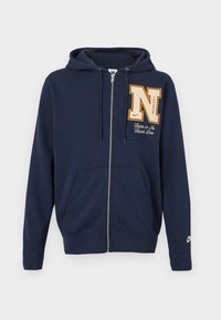 Tamsiai mėlynas džemperis su užtrauktuku, priekinėmis kišenėmis, didelė „N“ siūlė ant krūtinės su užrašu „There is No Finish Line“ ir mažu „Nike“ logotipu ant rankovės.