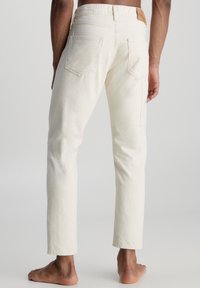 Jeans de mezclilla blanca con corte recto, que incluyen bolsillos traseros y una etiqueta de cuero con el logo en la cintura. Textura suave, diseño minimalista.