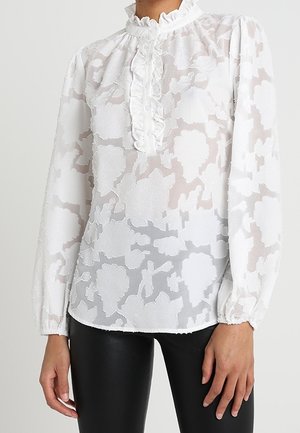 Biała przezroczysta bluzka z długim rękawem z kwiatowym wzorem żakardowym, z falbaniastym wysokim kołnierzem i plisą na guziki, noszona z czarnymi spodniami.