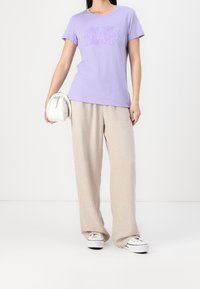 Levandulové bavlněné tričko s logem GAP, zkombinované s béžovými lněnými kalhotami a bílými teniskami. Nosí malou bílou kabelku.