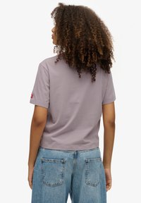 Camiseta morada de manga corta con corte recto, combinada con unos vaqueros de tiro alto en azul claro. Lleva una pequeña etiqueta de marca en la manga.
