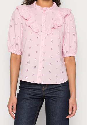 Vrouw draagt een lichtroze blouse met knopen, een ajour bloemenpatroon en een geplooide kraag en mouwen, gecombineerd met donkerblauwe spijkerbroek.