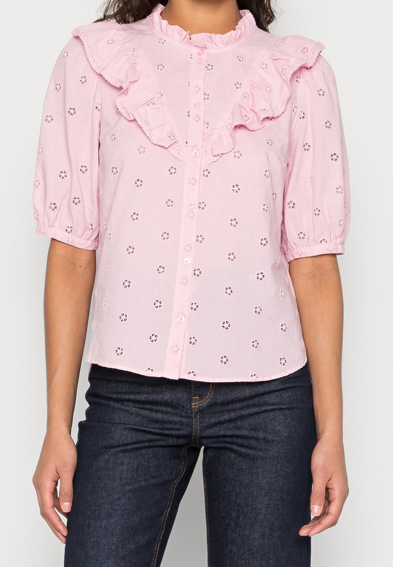 Une femme porte un chemisier rose clair boutonné avec un motif floral en broderie anglaise et un col et des manches à volants, associé à un jean bleu foncé.