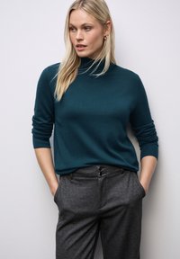 Teal, lange mouwtrui met een hoge hals, gecombineerd met grijze, op maat gemaakte broeken met een dubbelknoopsluiting en zijzakken.