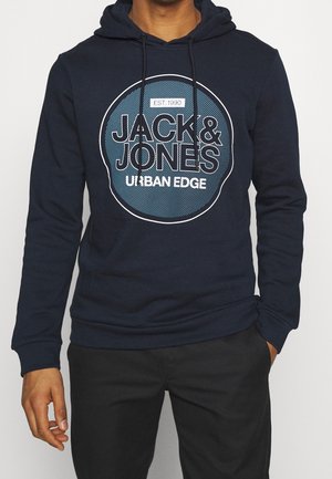 Mann trägt dunkelblauen Hoodie mit dem Logo "JACK & JONES URBAN EDGE EST. 1990" in einem Kreis, kombiniert mit schwarzer Hose, steht mit entspannten Händen.