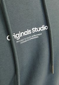 Dark green hoodie with a soft texture, featuring a white printed logo "Originals Studio" and silver accents on the drawstring.