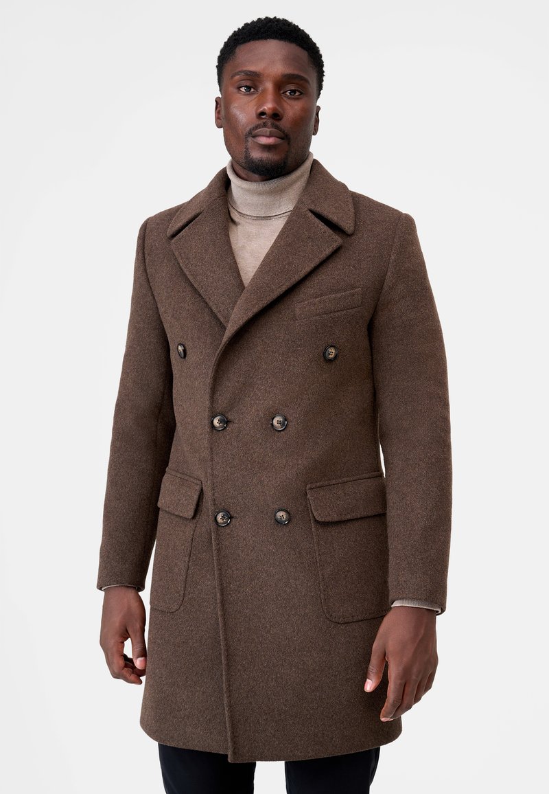 Brown wool double-breasted coat featuring black buttons, large front pockets, and a pointed collar. Worn over a beige turtleneck.
