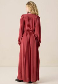 Femme aux cheveux blonds ondulés portant une longue robe rouge rouille avec une taille froncée et des manches longues ballons, se tenant de dos sur un fond neutre.