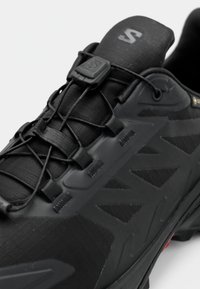Čierna športová obuv s rýchloupínacími šnúrkami a zosilneným textúrovaným zvrškom, navrhnutá na pou žitie vonku alebo na turistiku.
