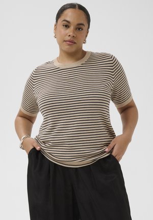Woman with dark hair in low bun wearing beige and black striped short-sleeve top and black pants, hands in pockets, neutral background.