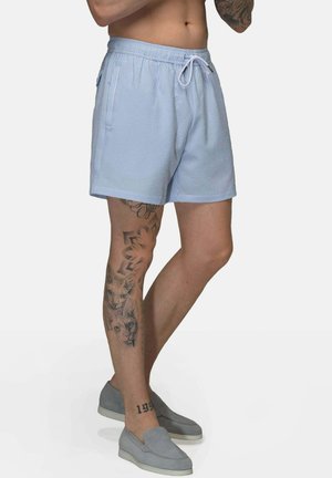 Mann trägt hellblaue Shorts mit Kordelzug und graue Slip-On-Schuhe, zeigt Tattoos am linken Bein und Arm vor einfarbigem Hintergrund.