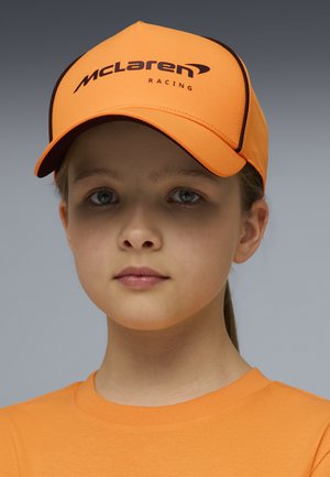 Giovane con occhi azzurri che indossa un cappellino arancione di McLaren Racing e una maglietta arancione abbinata, sfondo grigio neutro.