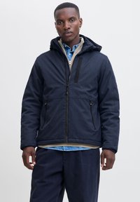 Marineblaues Jacket aus strukturiertem Stoff, mit Reißverschluss vorne und Kapuze. Verfügt über Fronttaschen und ein kontrastierendes Innenfutter. Kombiniert mit einem blauen Hemd.