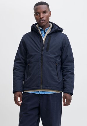 Chaqueta de marina con una tela texturizada, cierre frontal con cremallera y capucha. Presenta bolsillos delanteros y un forro interior en contraste. Combinada con una camisa azul.