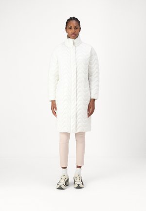 Witte gewatteerde jas met een hoge kraag, voorzien van een golvende textuur. Gecombineerd met lichtroze broeken en sneakers met zwarte accenten.