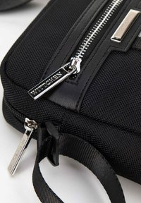 Borsa di stoffa nera con superficie texturizzata, cerniera e accessori in argento. Presenta una maniglia corta e un'etichetta con logo attaccata alla cerniera.