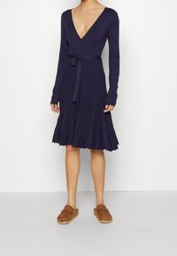 Mörkblå plisserad klänning med långa ärmar, djup V-ringning och knytband i midjan; den texturerade tyget skapar en svävande silhuett.