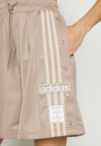 Beige idrottskjol med elastisk midja, tre vertikala vita ränder, tryckknappar på sidan och varumärkeslogotyper.