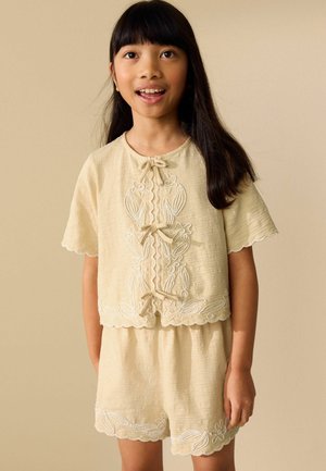 Young girl smiling, wearing a beige textured short-sleeve top and matching shorts with floral embroidery and scalloped edges.