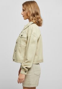 Chaqueta de mezclilla beige con mangas largas, cierre frontal de botones y bolsillos laterales. Diseño corto con mangas con puños y textura suave.
