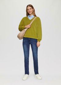 Grüner Pullover mit V-Ausschnitt, darunter ein blauer Hemdkragen, kombiniert mit dunklen Jeans und weißen Sneakers. Neutrales Detail der Umhängetasche.