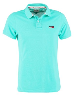 Man met een grijze bomberjack, turquoise poloshirt, donkere jeans en bordeauxrode sneakers staat tegen een effen lichte achtergrond.