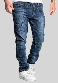 Denim jeans in dark blue with multiple stitched panels, a slim fit design, and various pockets, paired with white sneakers.