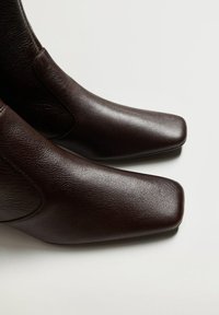 Botas de tobillo de cuero marrón con una textura suave, punta cuadrada y paneles laterales elásticos. El diseño es elegante con un estilo minimalista.