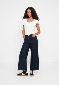 Jean en denim foncé à jambes larges avec une taille haute, associé à un haut ajusté à manches courtes blanc. Des baskets noir et blanc complètent le look.