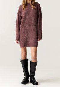 Robe en maille surdimensionnée couleur mauve avec ourlet et poignets côtelés, associée à des bottes en cuir noir brillantes et hautes. Texture lisse, coupe décontractée.