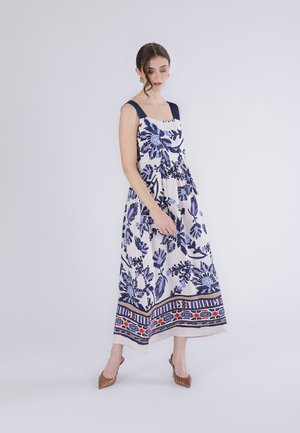 Nainen, joka pukeutunut hihattomaan valkoiseen mekkoon, jossa sinisiä kukkakuoseja ja koristeellinen reunus, seisoo hieman päätään kumartaneena yksinkertaista taustaa vasten.