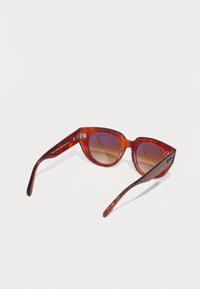 Gafas de sol de carey rojo con forma de ojo de gato sobredimensionada, lentes marrones con gradiente y detalles metálicos en las patillas. Acabado brillante.