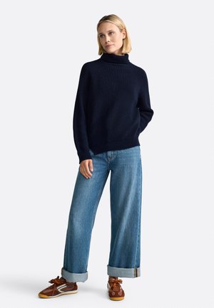 Granatowy sweter z prążkowanym golfem w połączeniu z jasnoniebieskimi spodniami typu wide-leg oraz brązowymi trampkami z pomarańczowymi sznurowadłami. Luźny i swobodny fason.