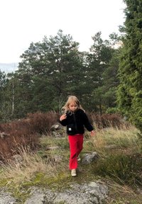 Schwarze Fleecejacke, rote Hose, auf steinigem Boden mit hohem Gras und Sträuchern im Hintergrund. Weiche Textur, lässige Passform, ohne Muster.