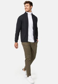 Sweat à zip gris foncé avec un col montant, porté sur un turtleneck blanc, associé à un pantalon vert olive clair et des baskets blanches.