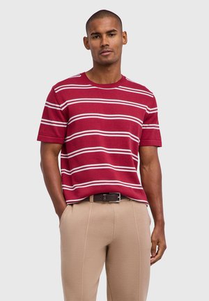 Mann trägt ein rotes Kurzarmhemd mit weißen horizontalen Streifen und beigefarbene Hose mit einem dunklen Gürtel, steht mit den Händen in den Taschen.