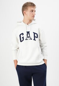 Kapuzen sweatshirt i krem med marineblå "GAP" logo, kengurulomme, hvite snorer og ribbestrikkede mansjetter. Sammen med marineblå bukser.