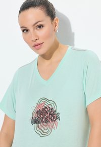 Mentolové tričko s výstřihem do V a krátkými rukávy, featuring dekorativní grafiku "Poutník v přírodě" v černé a červené barvě, obkreslenou zelenými tvary.