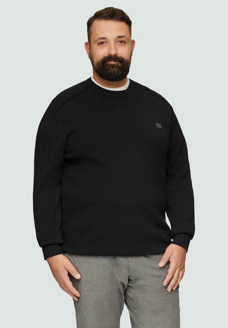 Schwarzer Strickpullover mit rundem Ausschnitt und gerippten Bündchen, versehen mit einem kleinen Logo auf der linken Brust. Kombiniert mit grauen strukturierten Hosen.