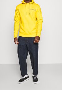 Felpa gialla con cappuccio e logo "HILFIGER", abbinata a jeans blu scuro a taglio largo e sneakers nere con lacci bianchi.