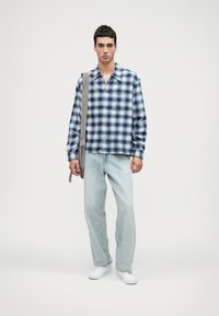 Πουκάμισο με κουμπιά σε μπλε και λευκή καρό σχέδιο, συνδυασμένο με ανοιχτό μπλε τζιν και λευκά αθλητικά παπούτσια. Το μοντέλο κρατάει μια μπεζ τσάντα ώμου.