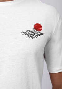 Λευκό βαμβακερό μπλουζάκι με μαύρο κλαδί με λευκά λουλούδια και έναν κόκκινο ήλιο γραφικά στην επάνω αριστερή πλευρά. Κοντά μανίκια.