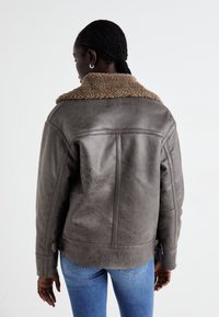 Brun läderjacka med en texturerad yta, prydd med en tanfärgad schäferkjortel och synlig sömnad. Modellen bär blå jeans.