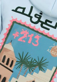 Lichtblauwe hoodie met een grafisch ontwerp van palmbomen, een moskee en de tekst "+213" in roze en Arabisch schrift.