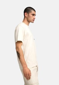 Camiseta de algodón beige con mangas cortas y cuello redondo, mostrada en un modelo de perfil. Presenta un logo simple en la parte frontal.