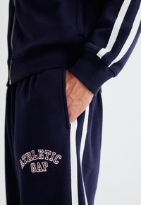 Marineblå treningsdrakt med hvite striper, inneholdende en jakke med glidelås og joggebukser med "ATHLETIC GAP" brodert, myk tekstur, skrå lommer.