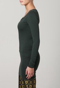 Tmavě zelený top s dlouhými rukávy a designem s V výstřihem, vyrobený z měkké tkaniny, má přiléhavý siluetu a hladkou strukturu.