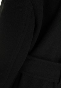 Czarny wełniany płaszcz o teksturowanym wykończeniu, z paskiem w talii i usztywnionymi rękawami. Tkanina wydaje się miękka i gęsta.